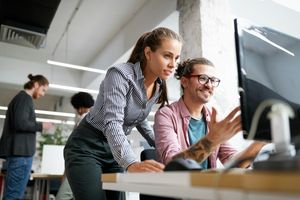 Two coworkers collaborate at a computer in a modern office, with a woman leaning in to look at the screen while a man gestures and smiles.